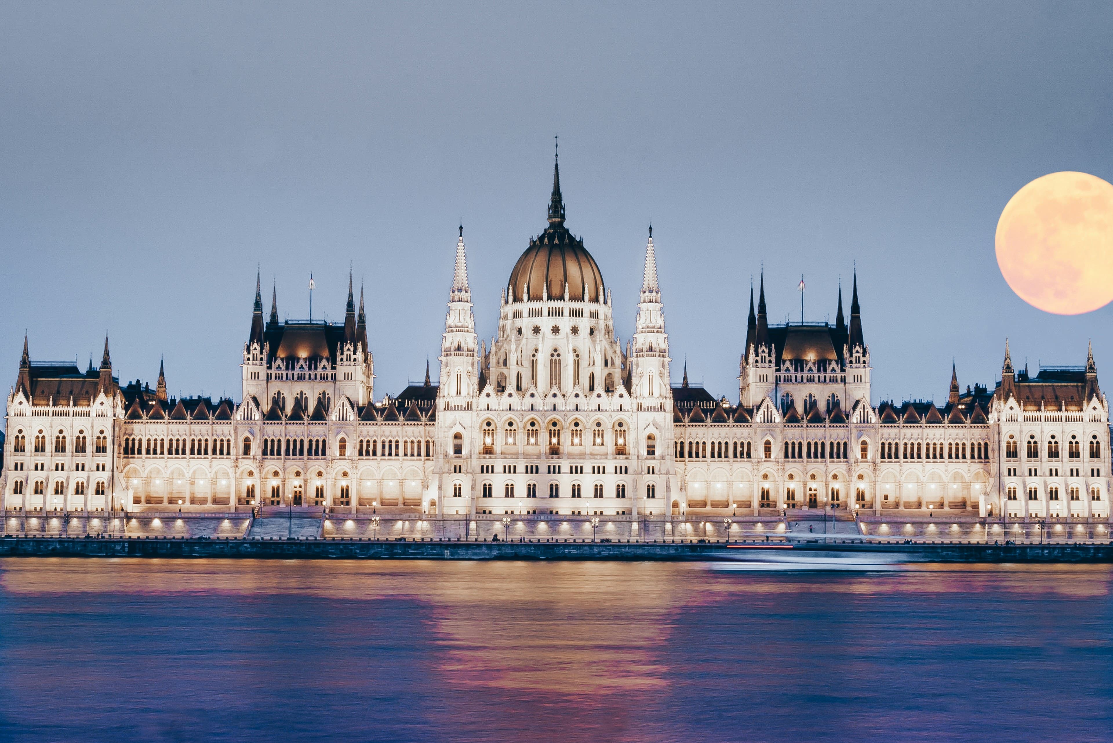 The Hungarian Parliament Building lit up at dusk on the Danube, a full moon rising beside its dome.