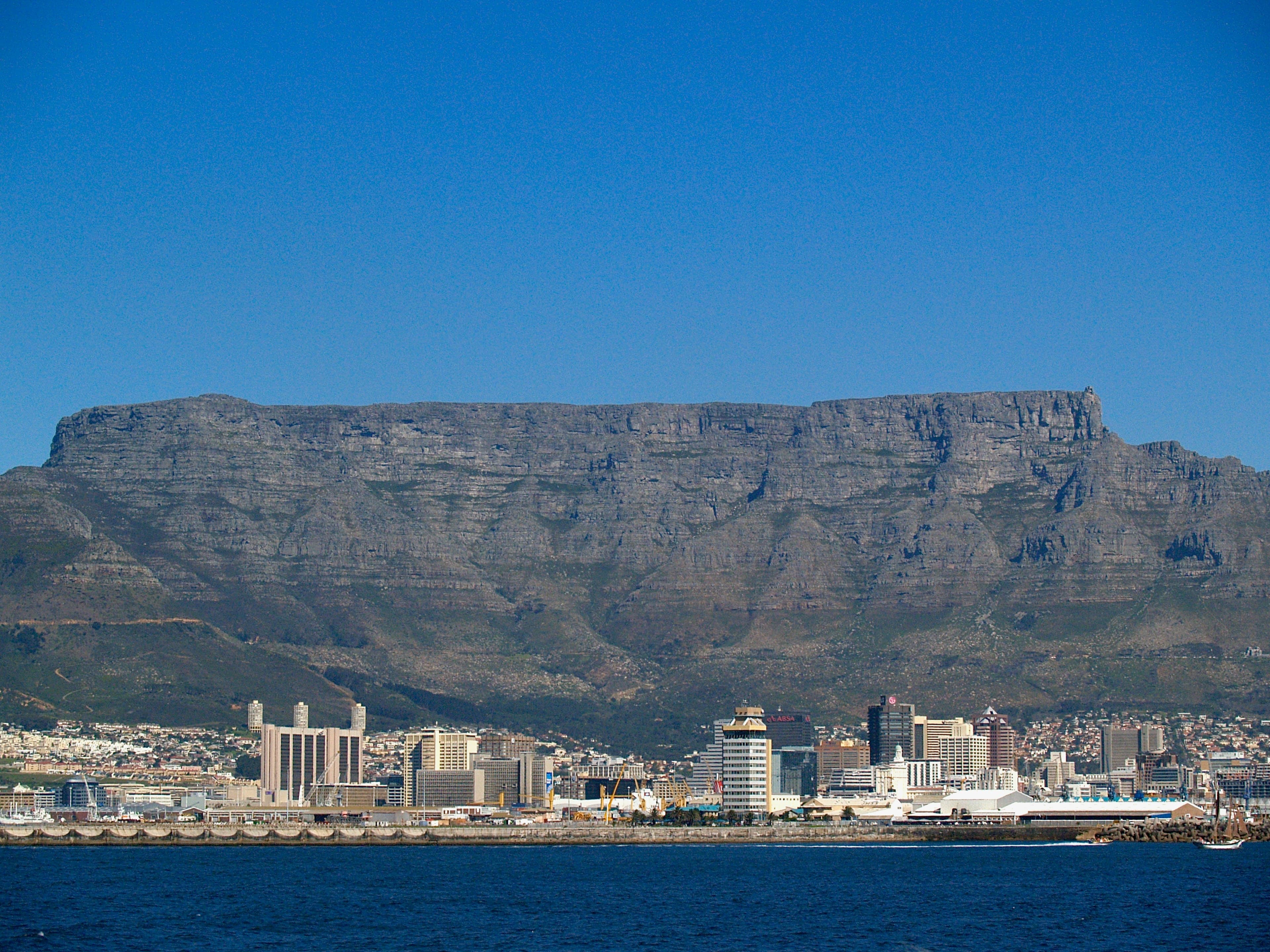 Table Mountain rising over Cape Town — its flat summit above the city skyline and harbour, under a clear blue sky with the sea in the foreground.