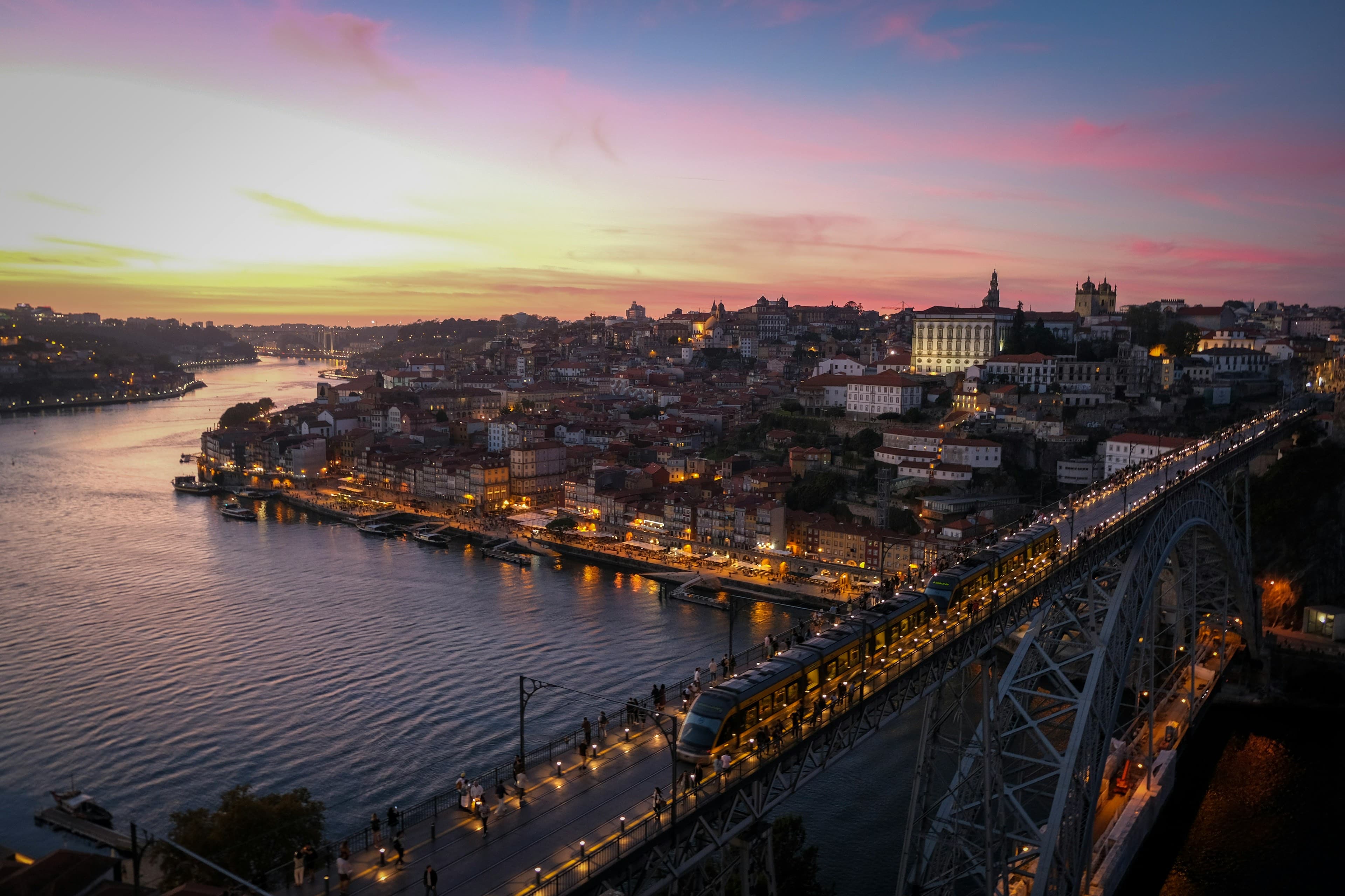The Dom Luís I bridge over the Douro at dusk, a tram crossing above Porto's lit-up Ribeira riverfront under a pink sunset.