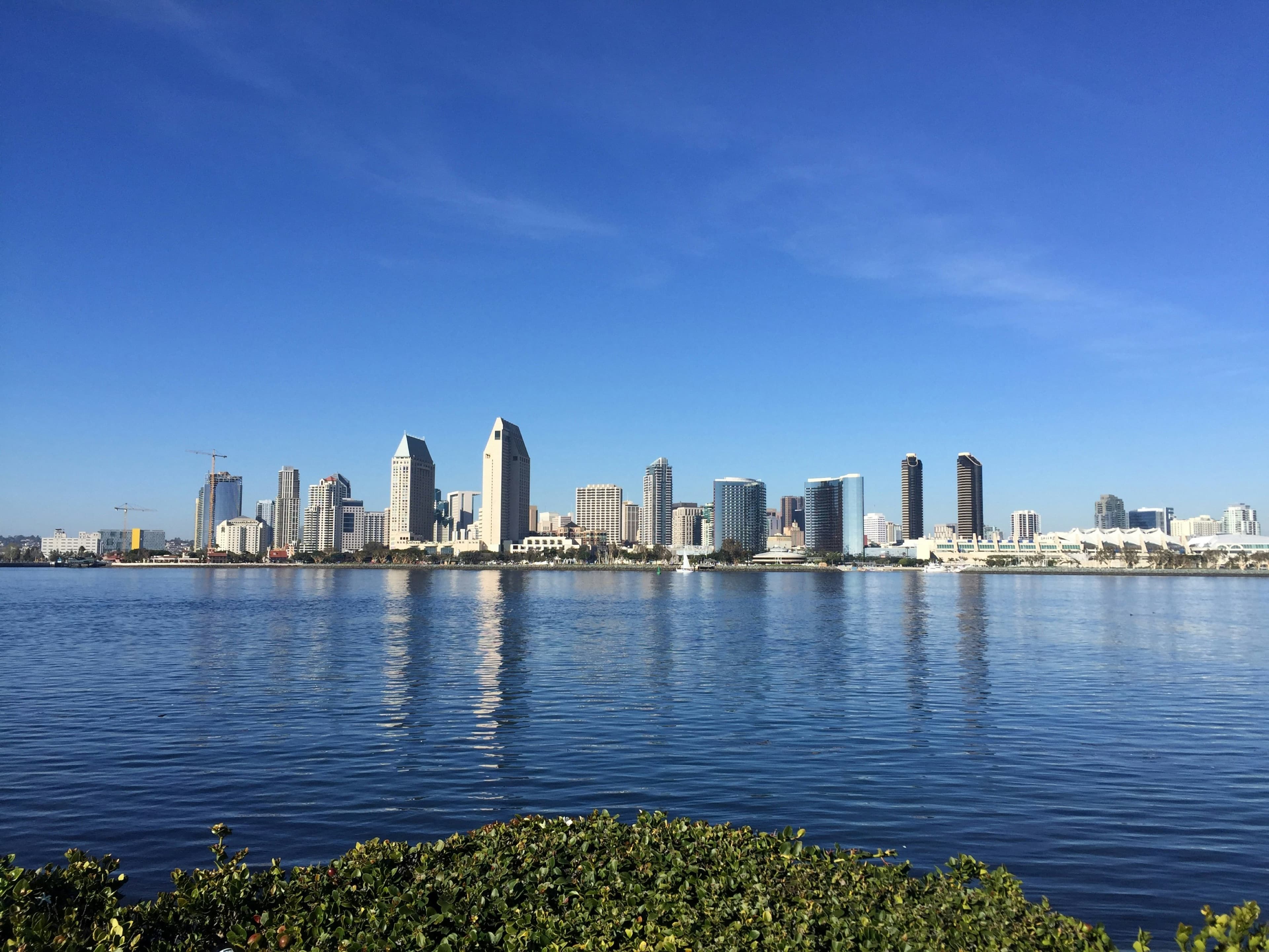 The downtown San Diego skyline across the bay from Coronado under a deep blue sky, the waterfront reflected in the water.