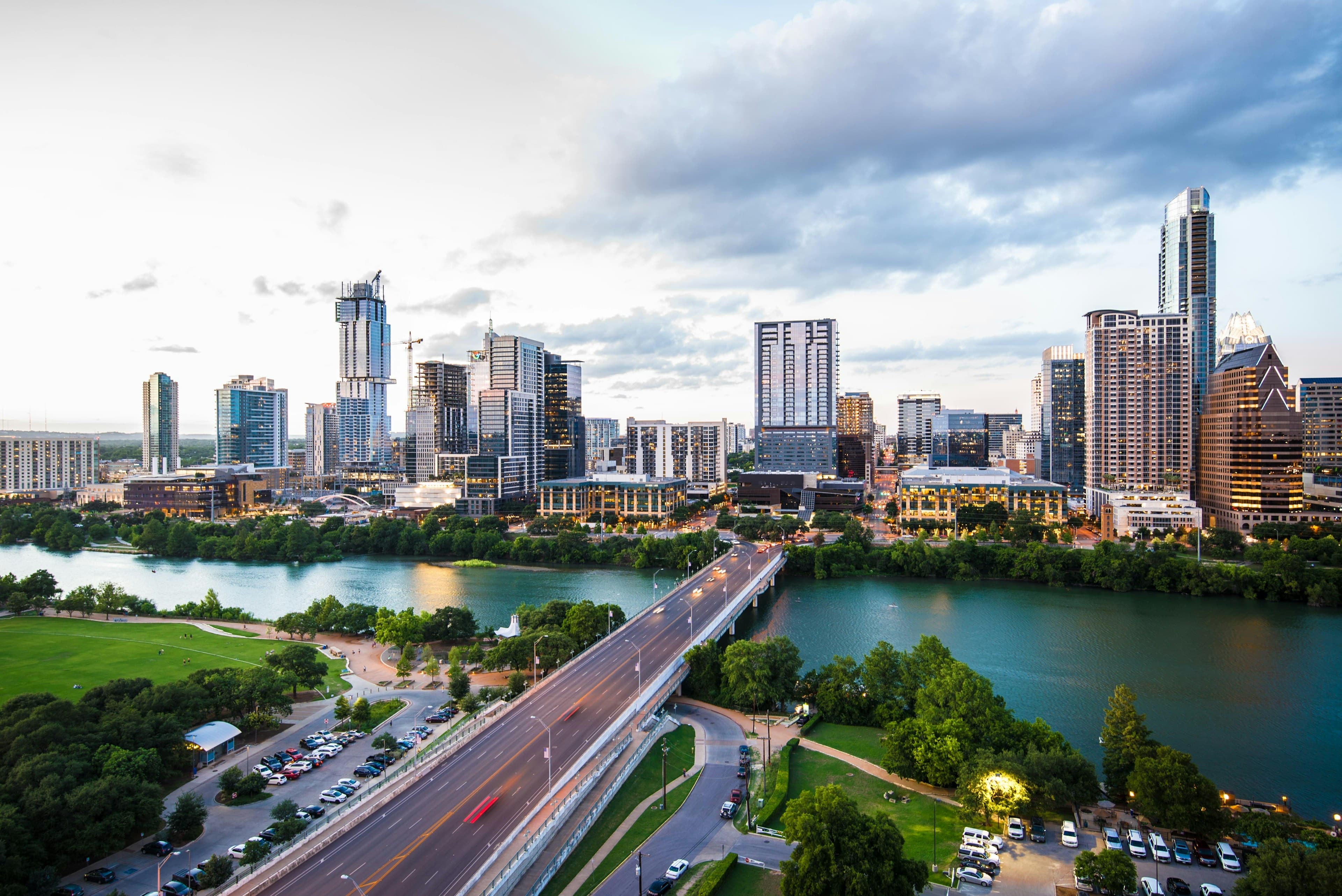 The downtown Austin skyline at dusk above Lady Bird Lake, the Congress Avenue bridge crossing the water with green parkland on both banks.