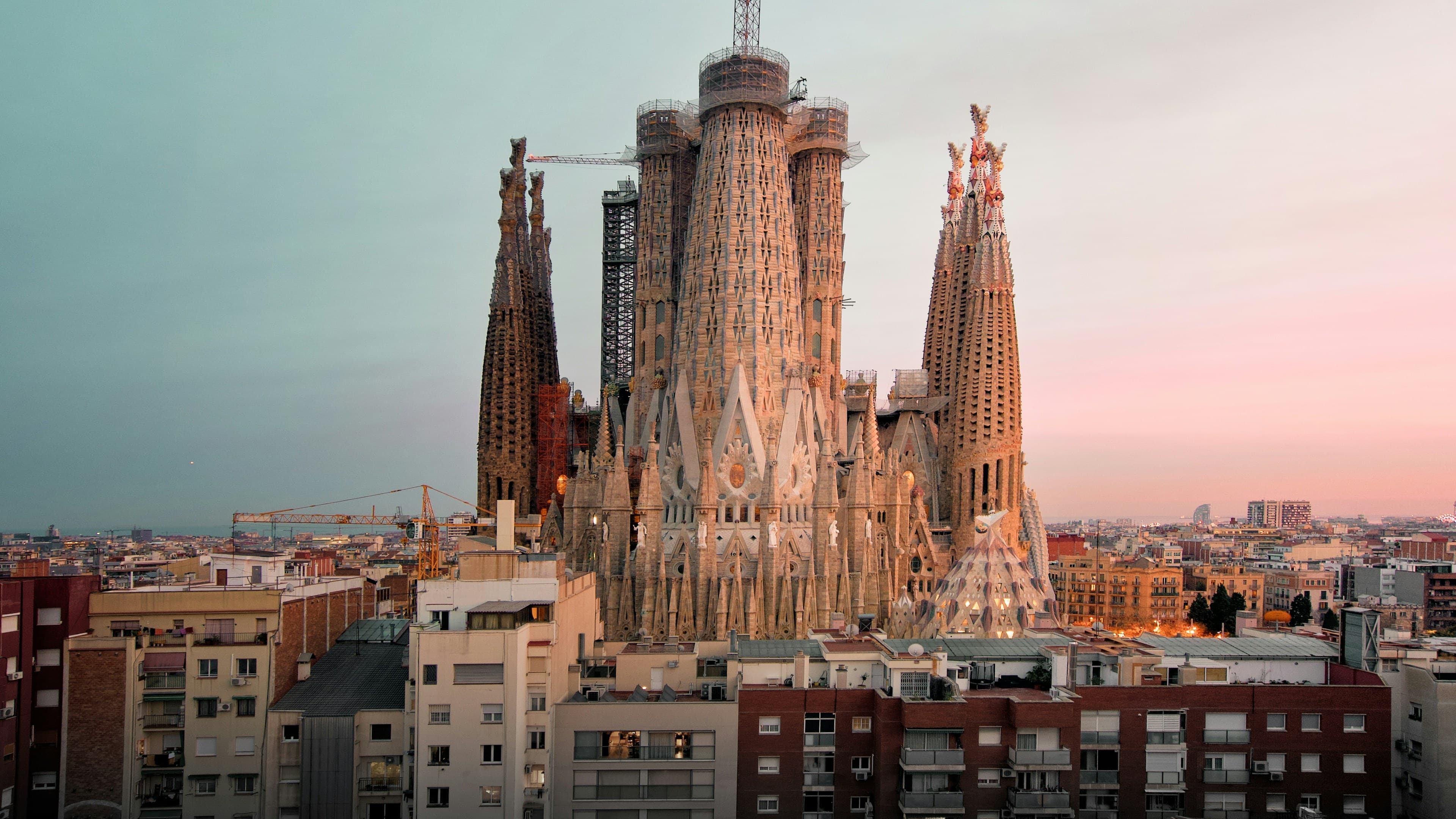 The Sagrada Família rising above Barcelona's rooftops at golden hour, the city stretching to the Mediterranean under a pink-and-teal sky.