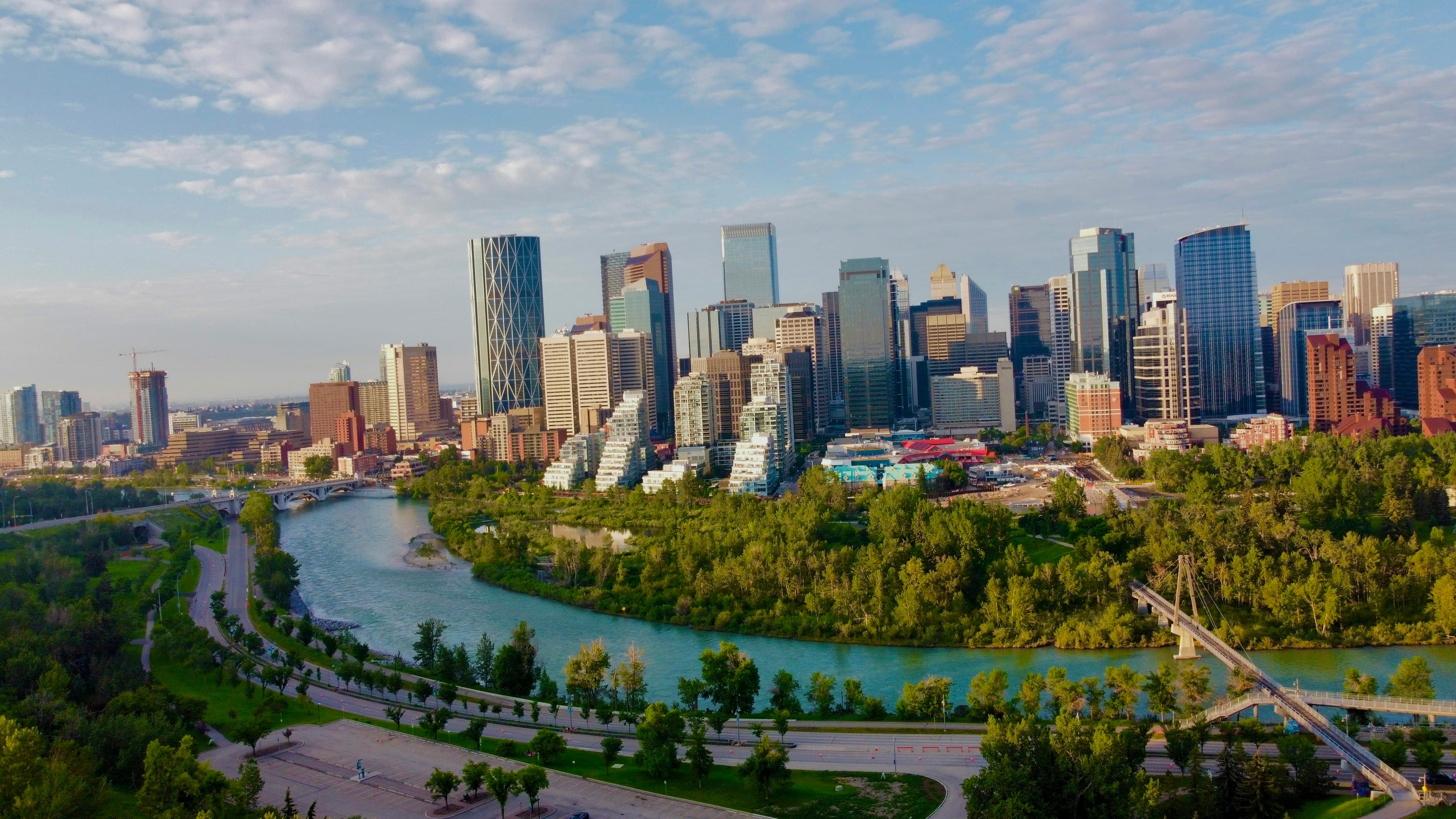 The downtown Calgary skyline above a bend in the turquoise Bow River, riverside parks and a footbridge in the foreground.