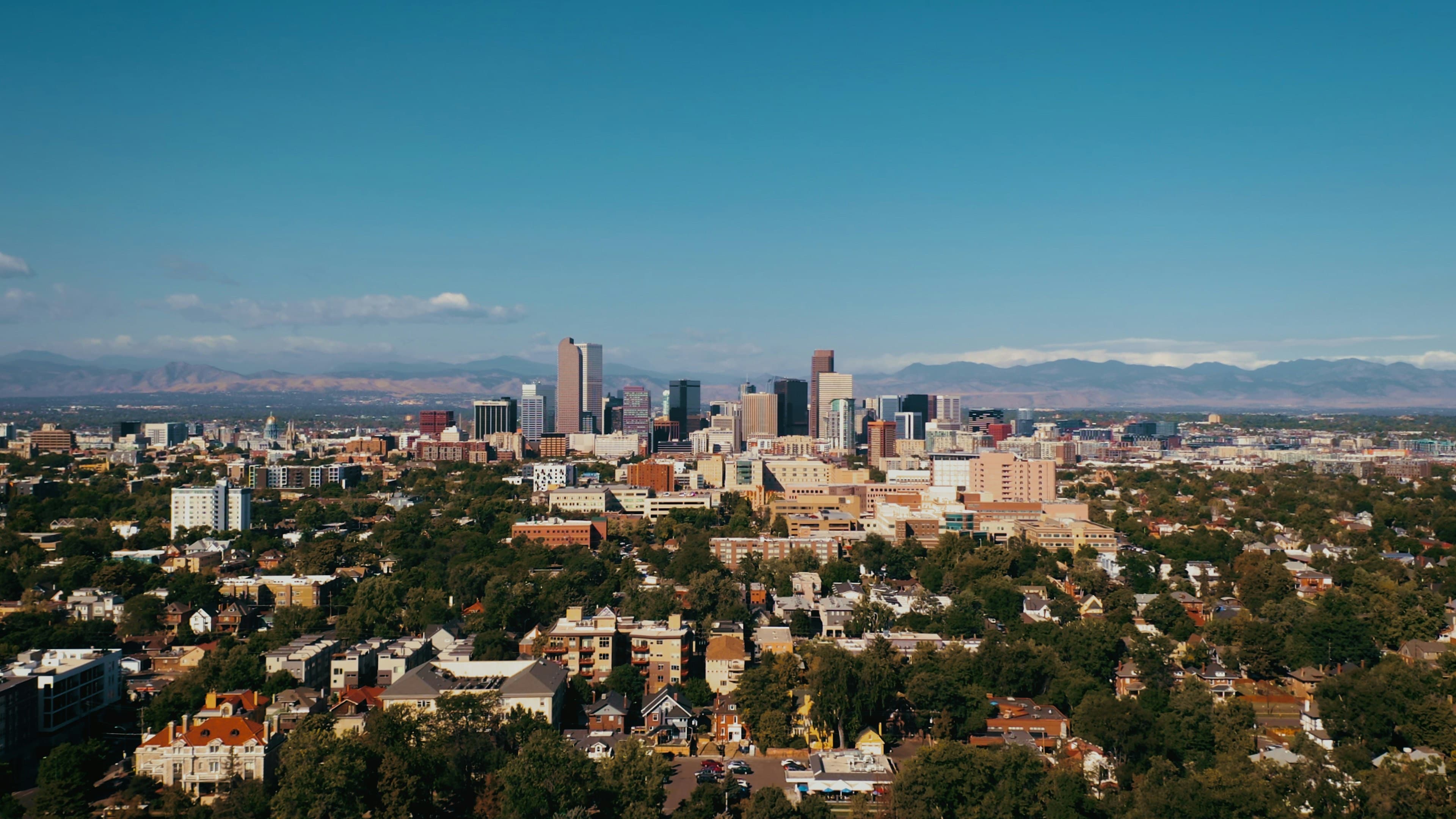The downtown Denver skyline under a blue sky with the Rocky Mountains on the horizon and tree-filled neighborhoods in the foreground.