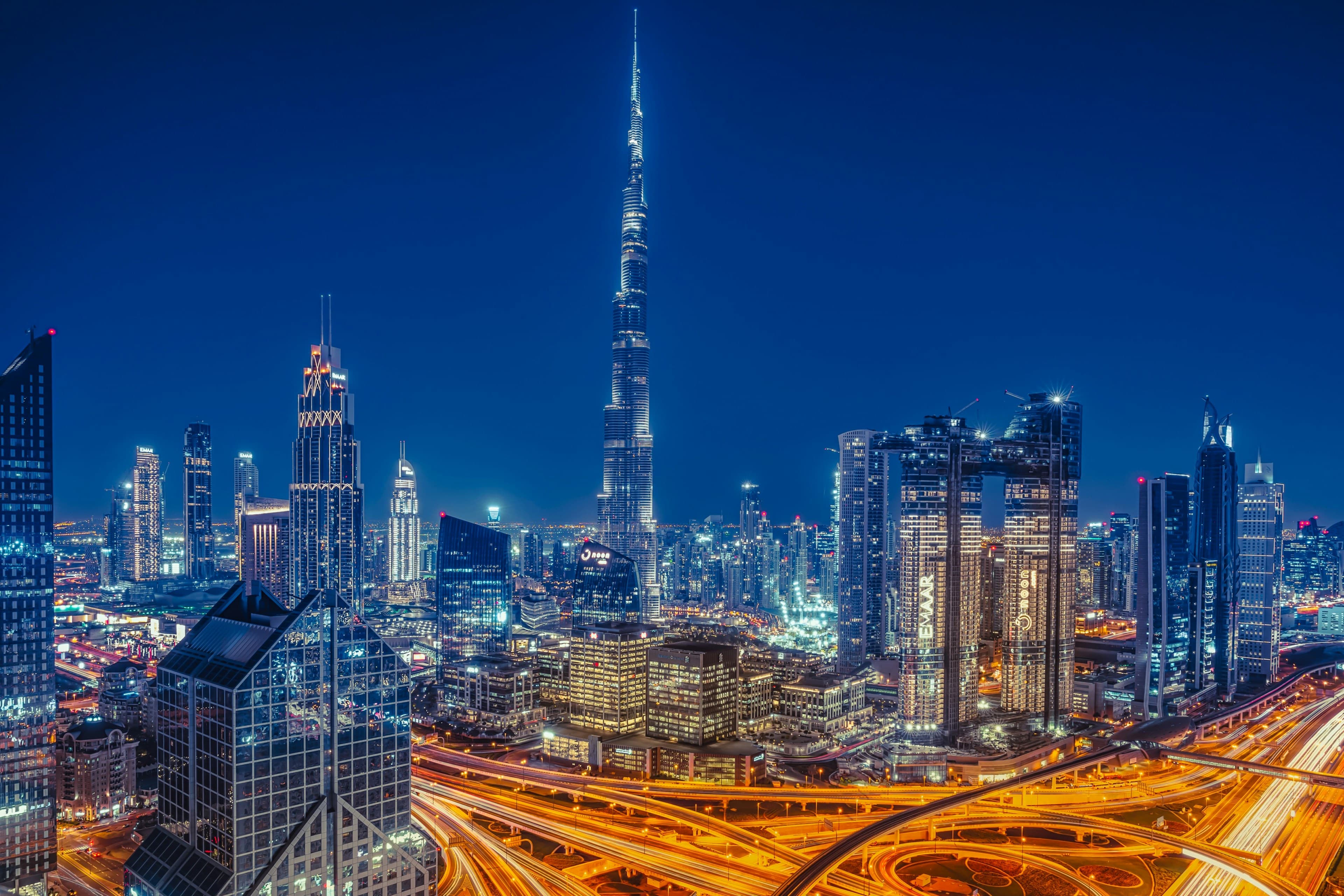 Downtown Dubai at blue hour — the Burj Khalifa towering over a glittering, lit-up skyline, with golden highway light trails sweeping across the interchange below.