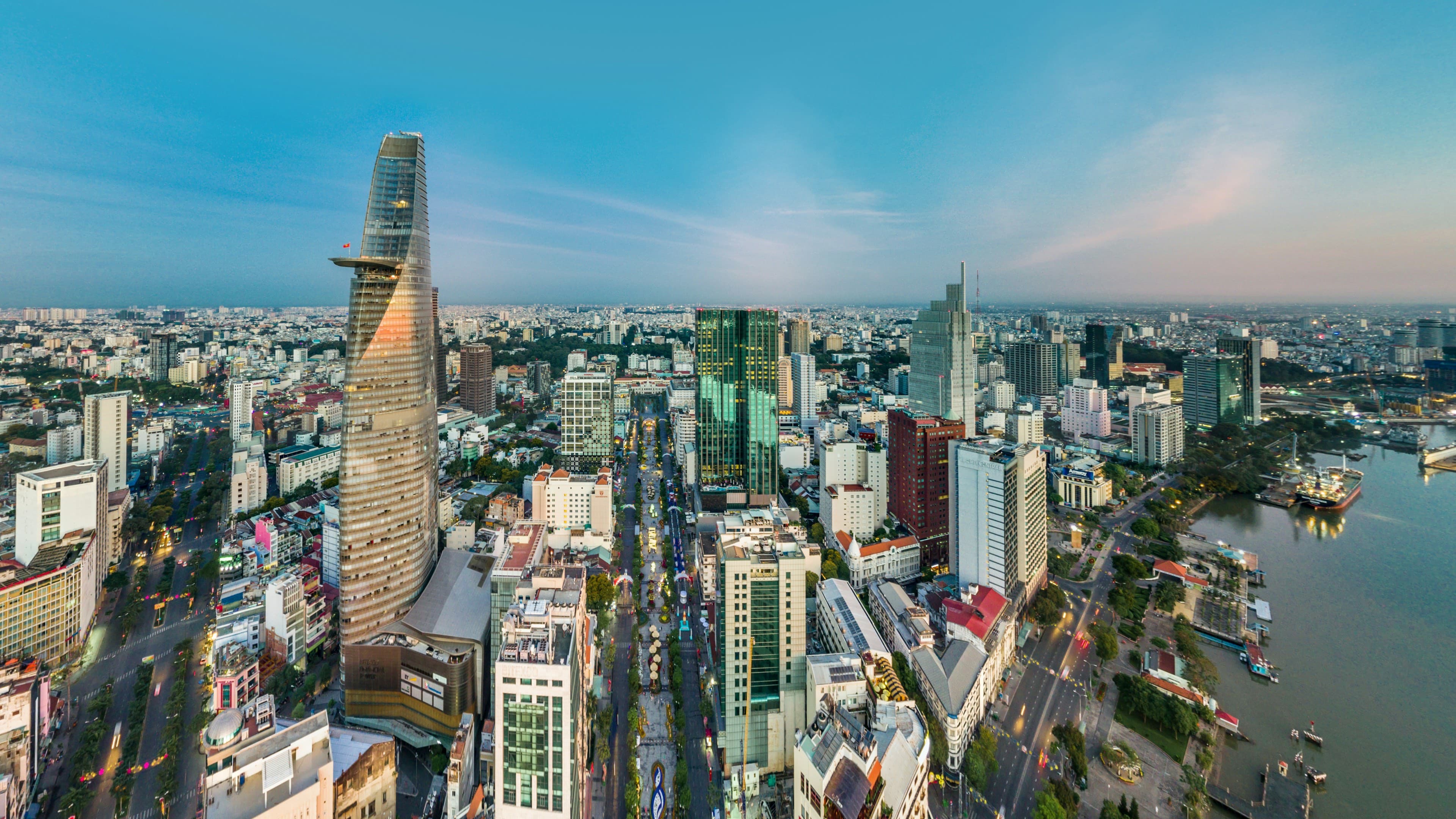 Aerial of downtown Ho Chi Minh City at dusk — the Bitexco Financial Tower over District 1 and the Nguyễn Huệ boulevard, the Saigon river beyond.