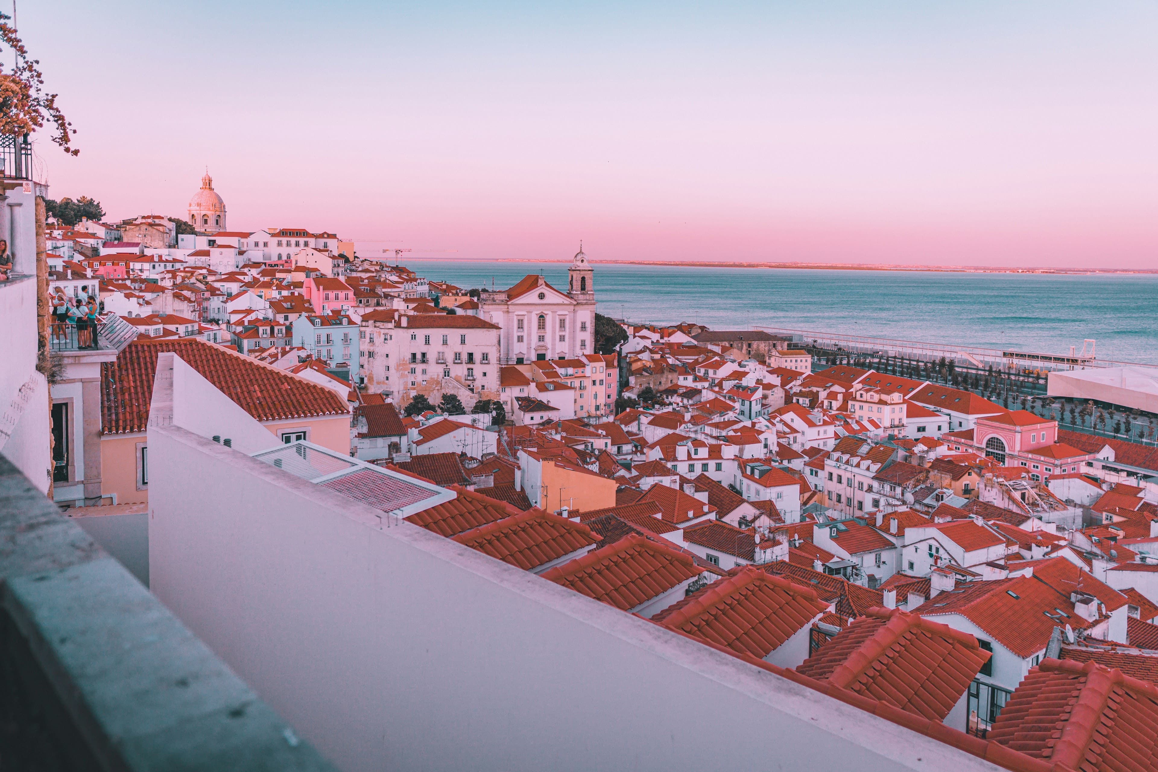 The Alfama district of Lisbon at pink sunset, terracotta rooftops sloping down to the Tagus with the National Pantheon dome above.