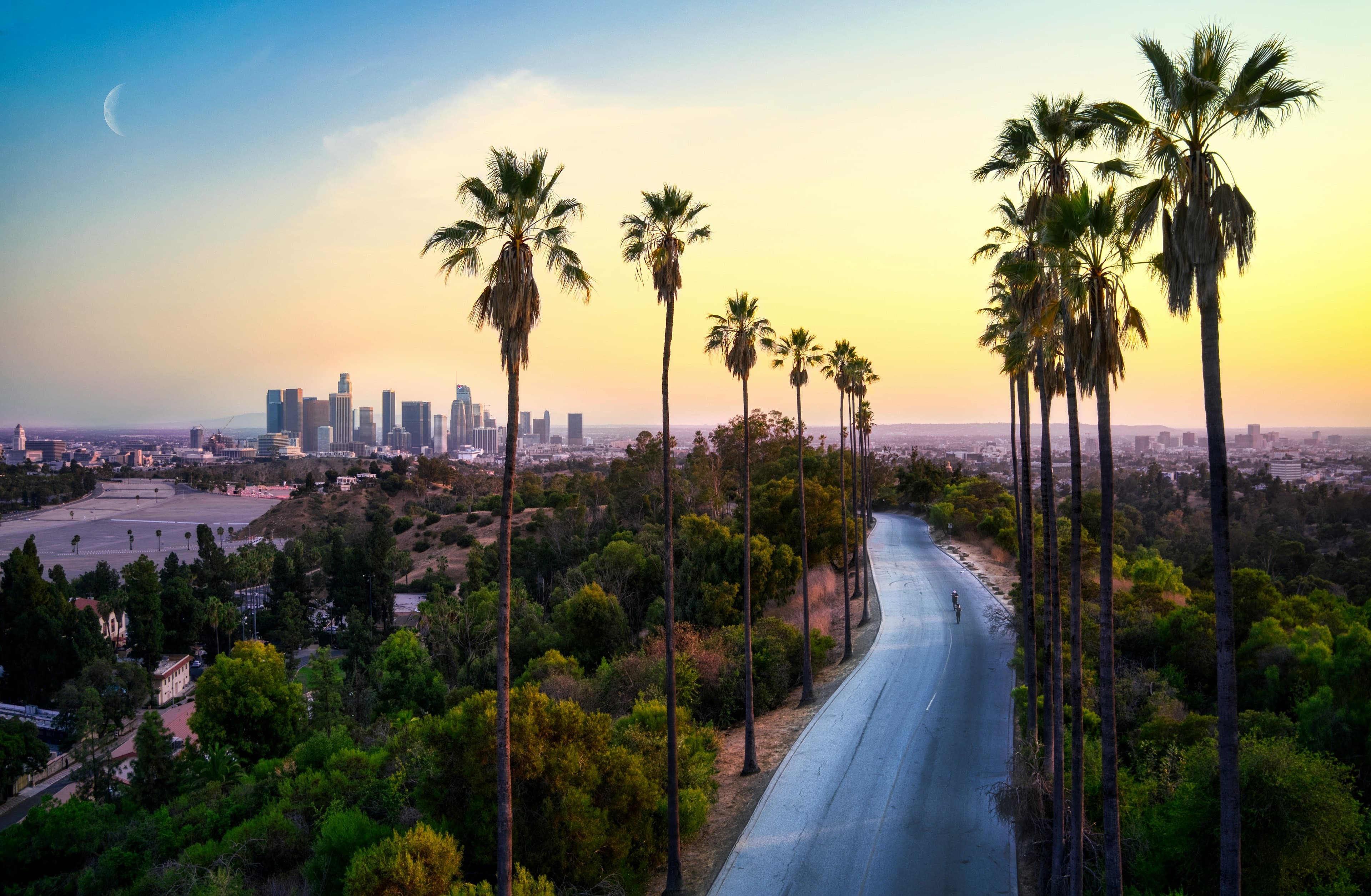 A palm-lined road curving toward the downtown Los Angeles skyline at golden-hour sunset, a crescent moon in the sky.