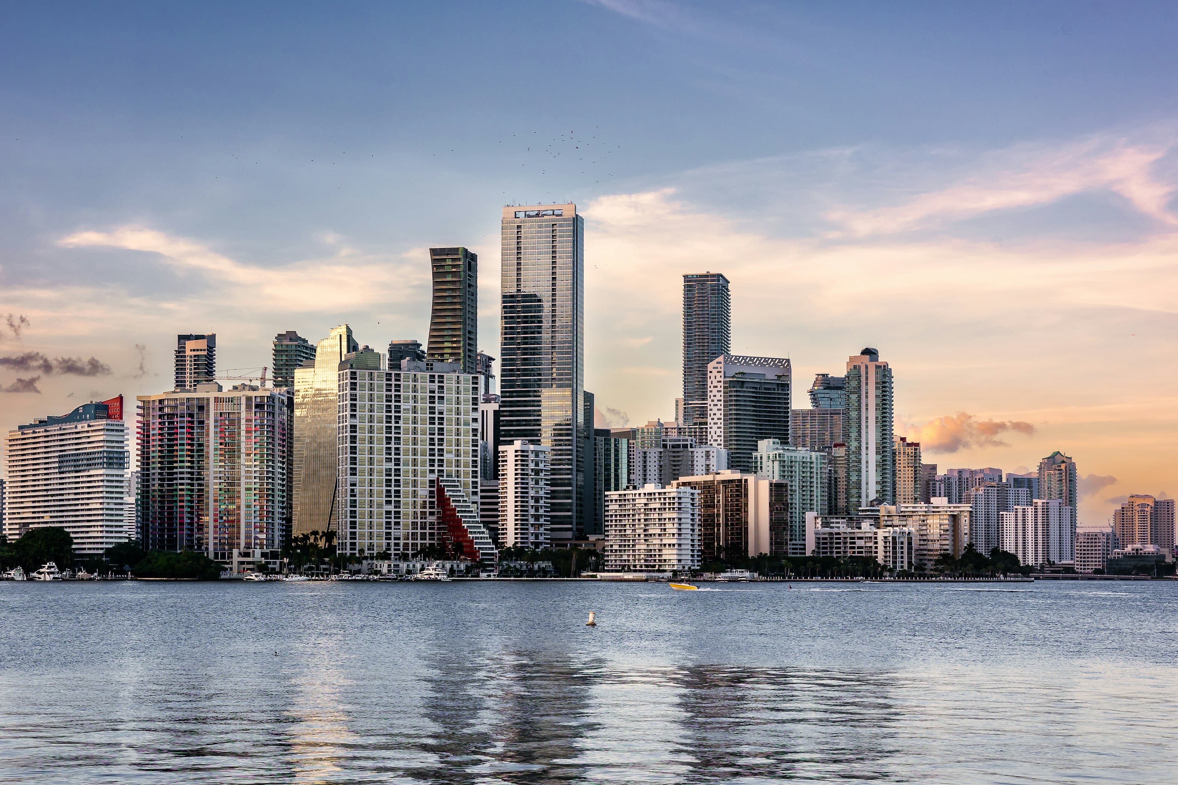 The Brickell skyline of Miami glowing at golden hour across Biscayne Bay, glass towers reflected in the calm water.
