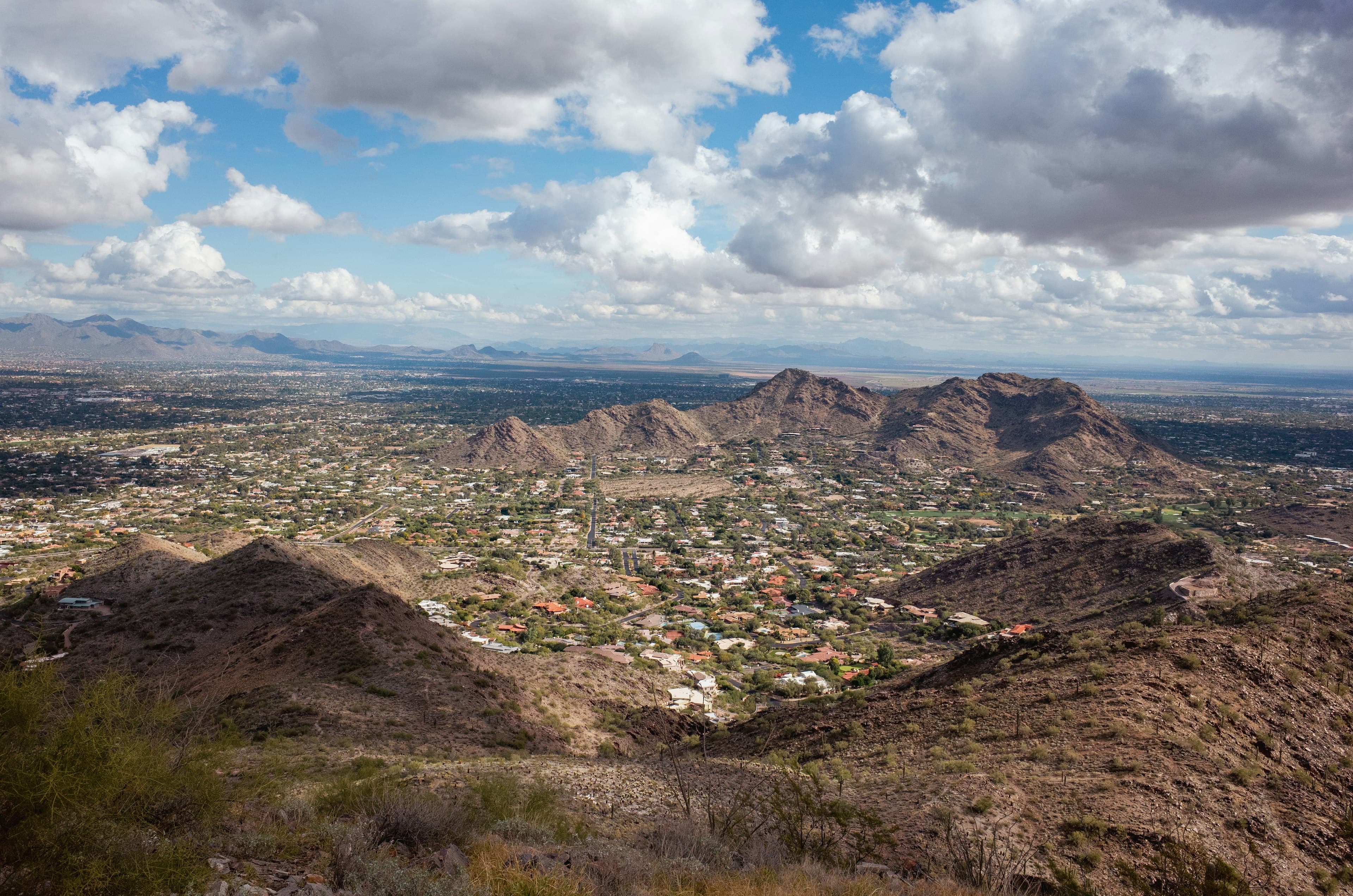 The Sonoran Desert around Scottsdale seen from a hilltop, desert mountains above the city spread across the valley under a big cloudy sky.