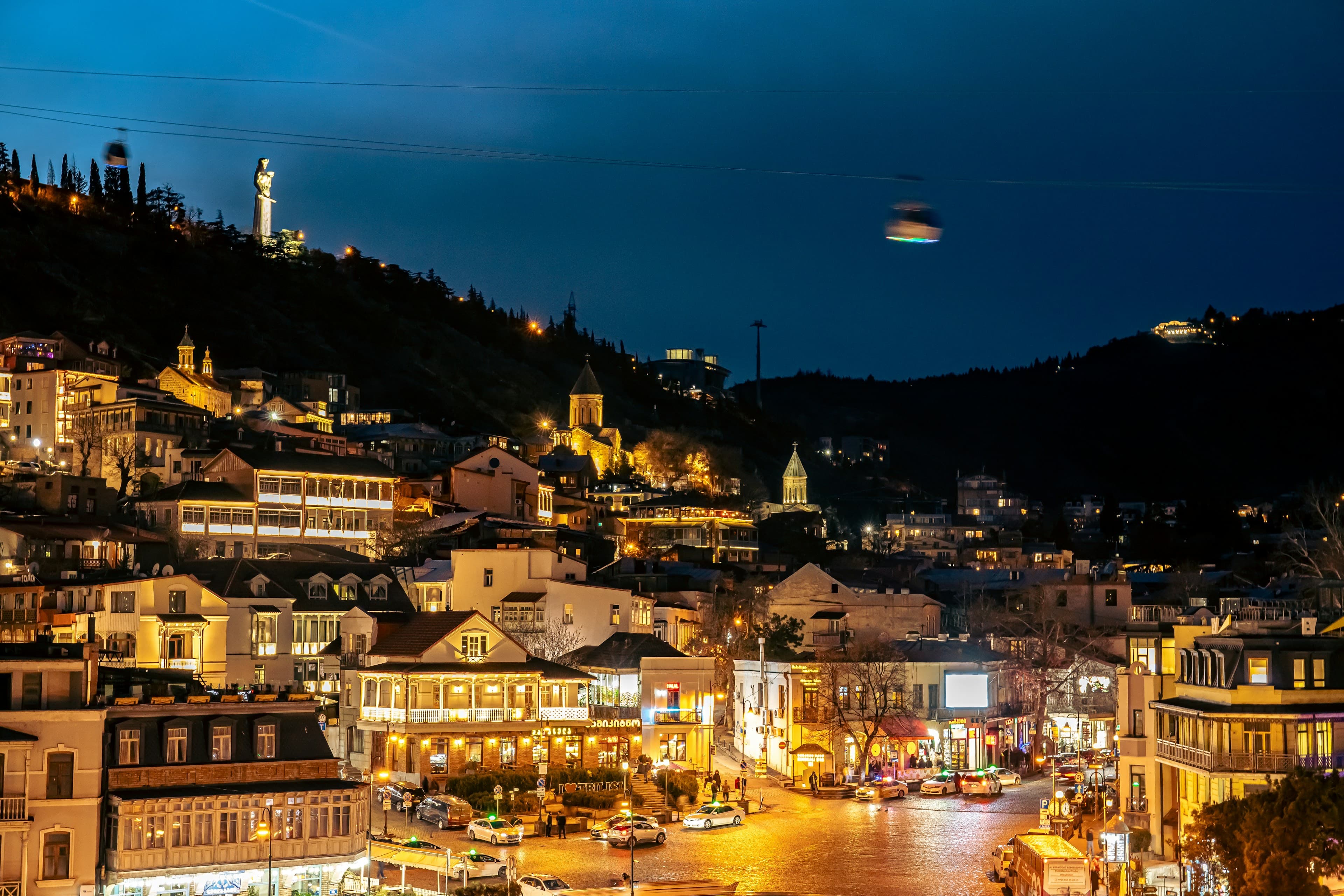 Tbilisi's old town at blue hour, wooden-balconied houses lit warm on the hillside below the Mother of Georgia statue.